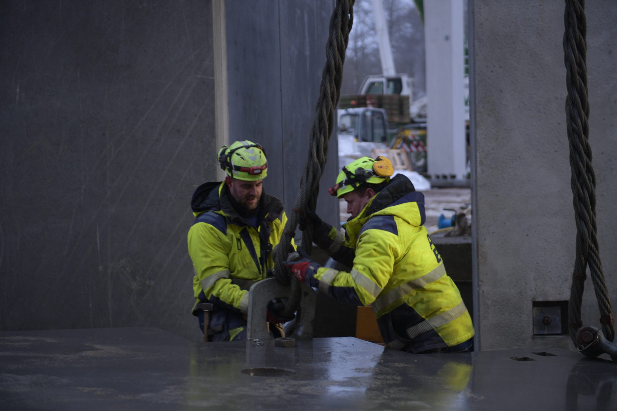 To BMS-riggere fastgør en løftewire til et betonelement på en byggeplads