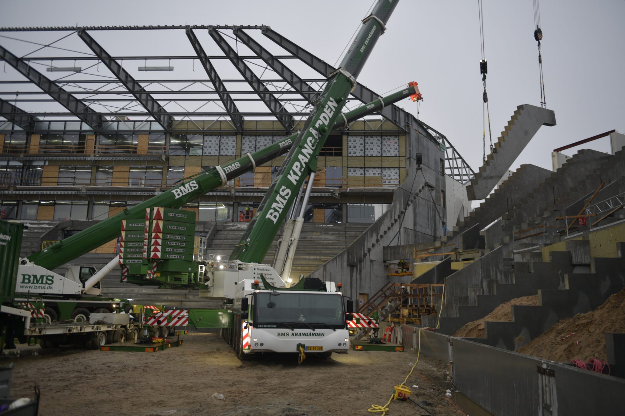 To BMS Krangården-kraner løfter et præfabrikeret betontrappeelement på plads under opførelse af et stadion