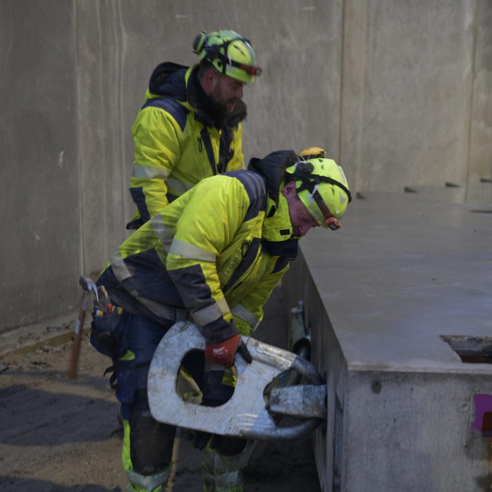 To BMS-riggere fastgør en krankrog til et betonelement på en byggeplads