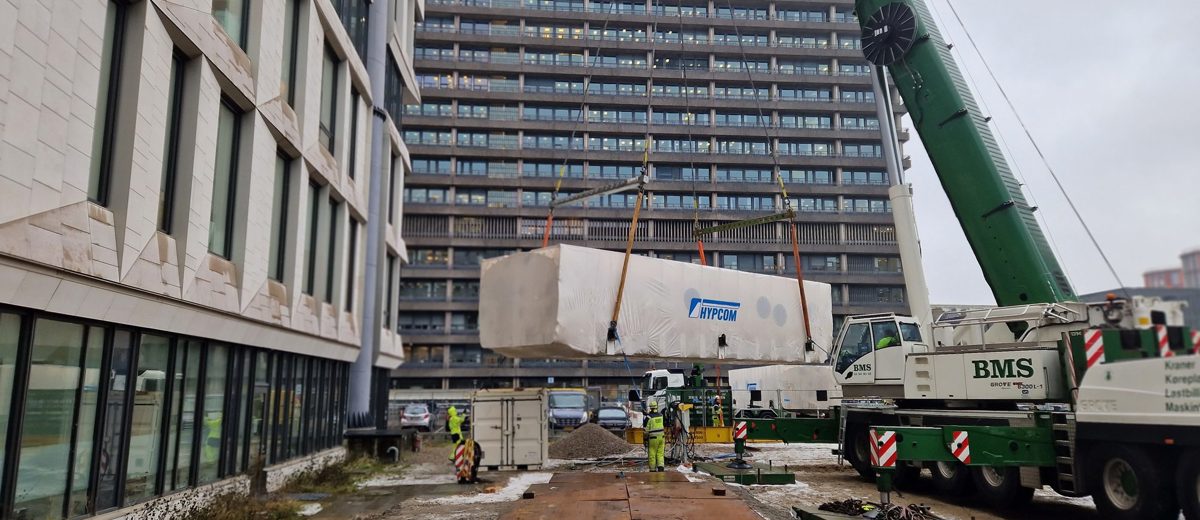 BMS-kran sænker et stort indpakket teknikmodul ned mellem bygninger ved rigshospitalet på køreplader